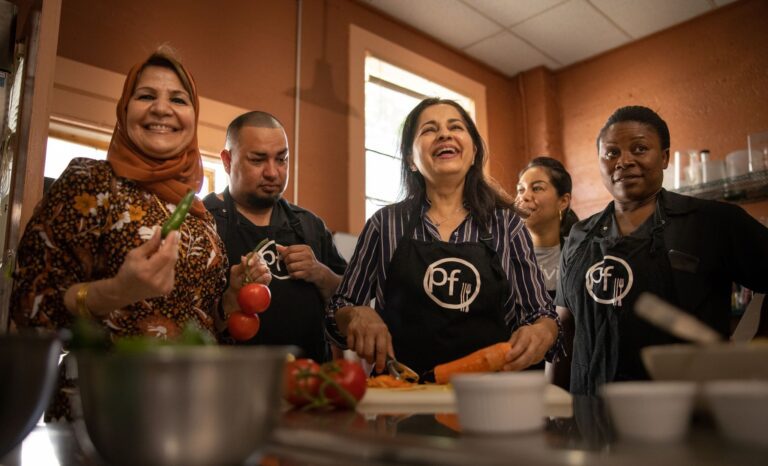 A smiling group of four present dishes they've cooked in a warmly lit kitchen