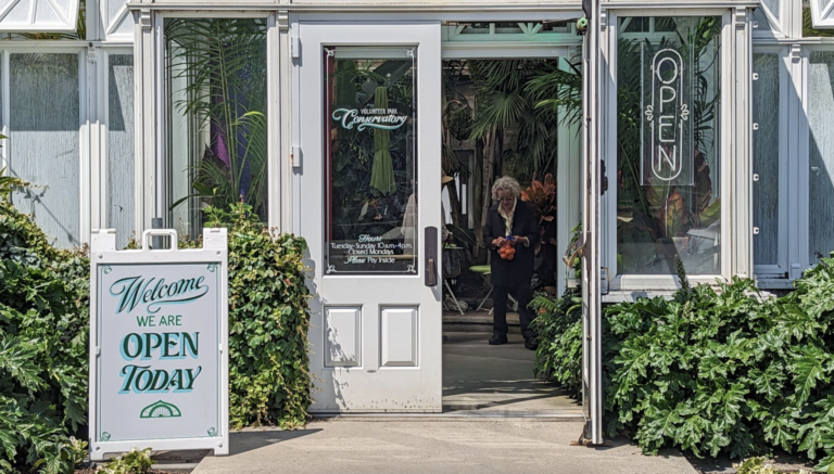 The entrance to the Volunteer Park Conservatory with the door open and a Come In We're Open sign out front.
