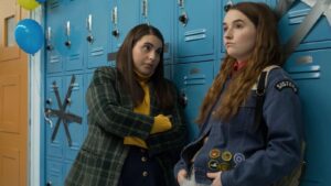 A girl looks sternly at another girl in a high school hallway in front of teal blue lockers. It is a still from the movie "Booksmart" which is screening soon at the SIFF Cinema Egyptian