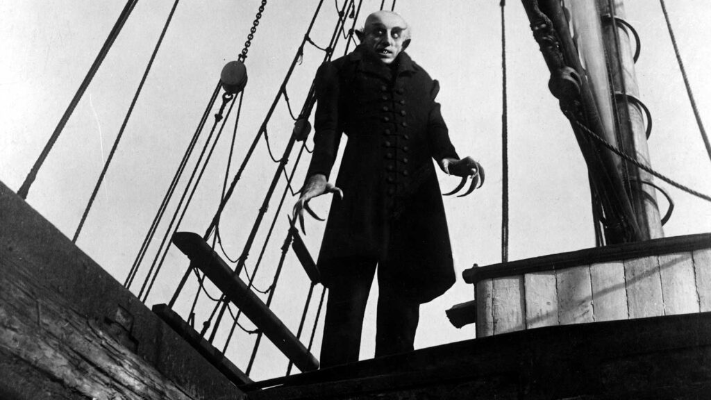 Nosferatu stands in the background, looking creepy, on a ship. An old black and white film still.