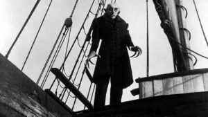 Nosferatu stands in the background, looking creepy, on a ship. An old black and white film still.