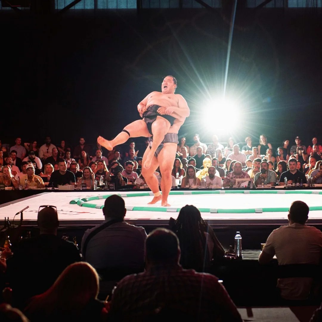 A sumo wrestler gets thrown while an attentive crowd watches.