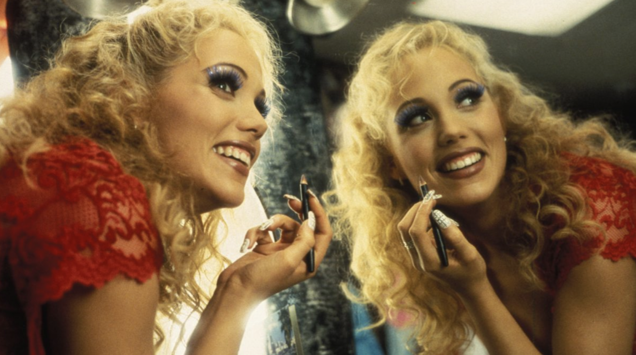 A still from the movie Showgirls featuring the character Nomi Malone looking away from a mirror while applying lipstick and smiling.