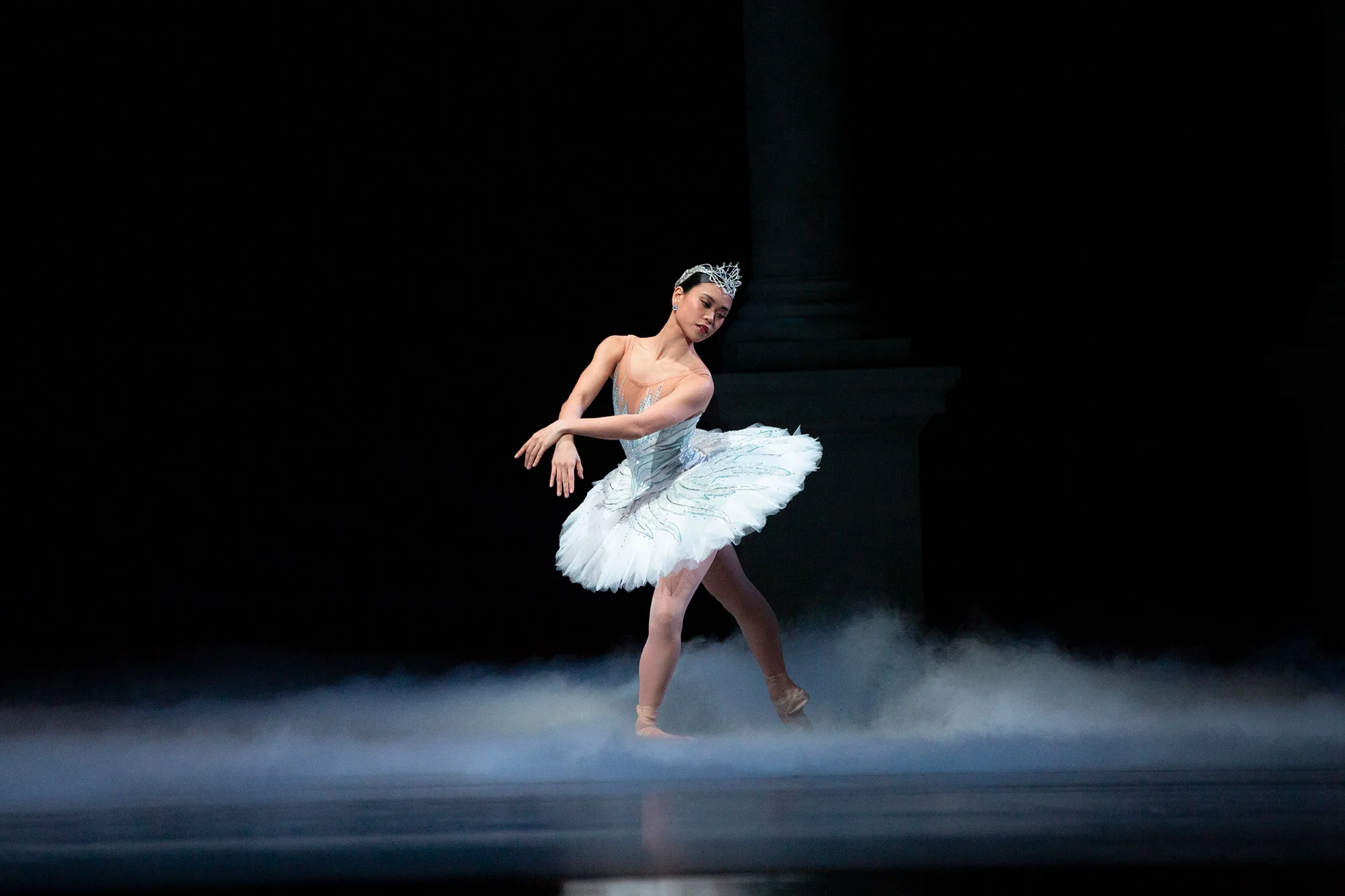A spotlit Angelica Generosa in Swan Lake holds a pose on a black stage with mist swirling around her feet.