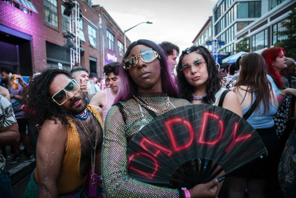 Three Queer/Pride partygoers in Seattle, Washington, pose for a picture in the Capitol Hill neighborhood, a historic queer neighborhood. The person in the middle holds a fan that says DADDY in red letters.