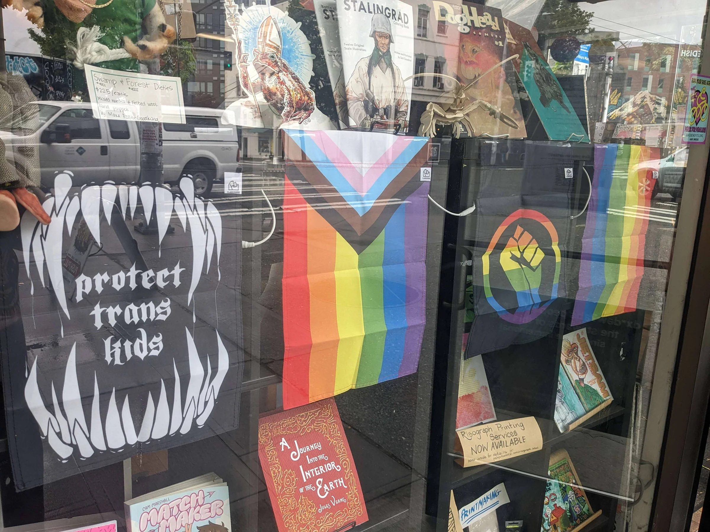 Push/Pull's window display features pride flags and social justice posters alongside indie comics and zines, with book covers and artwork arranged on multiple levels against a reflective glass storefront.