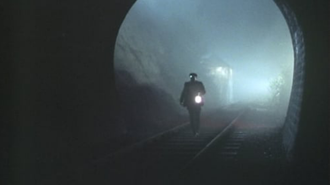 A still from BBC's Ghost Stories for Christmas series showing a solitary figure in Victorian dress walking away into a misty railway tunnel, holding a glowing lantern that casts an eerie light through the fog.