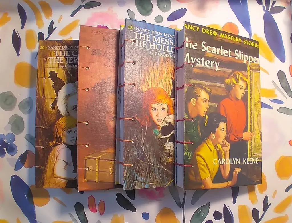 Four vintage Nancy Drew mysteries transformed into handbound journals with red stitching, displayed against a watercolor-splattered background. Book covers show the iconic mystery series' dramatic artwork featuring the teenage detective.