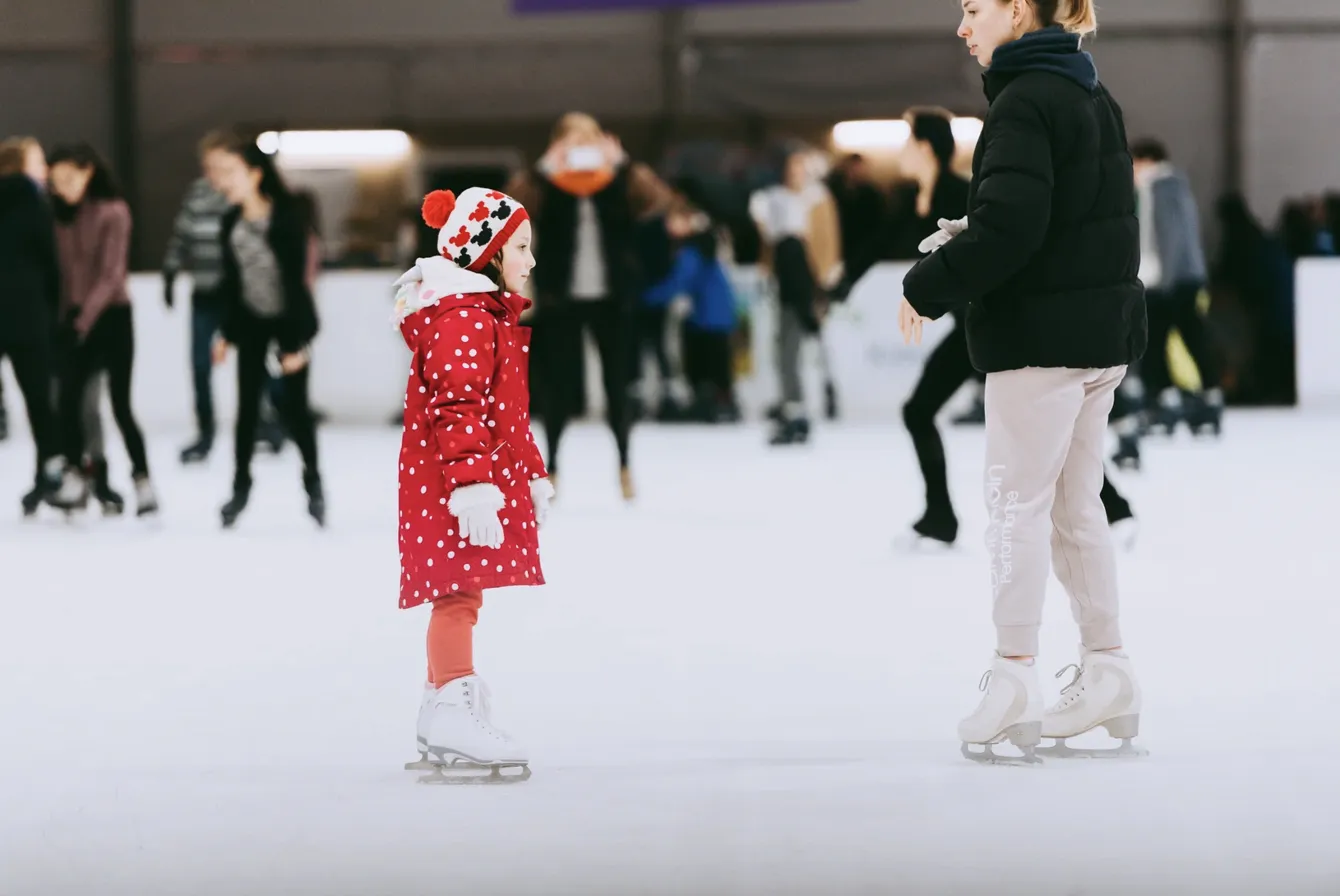 A young skater in a red polka dot winter coat and Mickey Mouse winter hat enjoys ice skating at an indoor rink in the Seattle area, including Bellevue. The bright, well-lit venue features a smooth ice surface where families and skaters of all skill levels can enjoy winter recreation. Professional white figure skates are visible in the foreground, while other skaters practice in the background, making this indoor winter activity accessible regardless of weather.