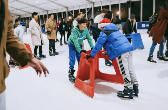 Two young skaters use a red training aid at an indoor ice rink during the holiday season, with one wearing a festive Santa hat. The skating assist device helps beginners learn to balance while other skaters practice in the background under twinkling lights in the covered venue. Many Seattle-area ice rinks provide these popular training aids for first-time skaters, making ice skating accessible for all skill levels.