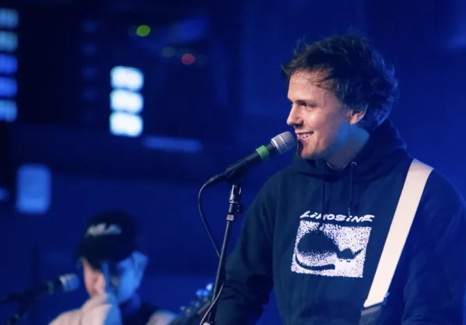 A singer-performer smiles while speaking into a microphone on stage, wearing a black hoodie with white graphic print, illuminated by blue stage lighting. Musical equipment and another performer are visible in the soft-focused background.