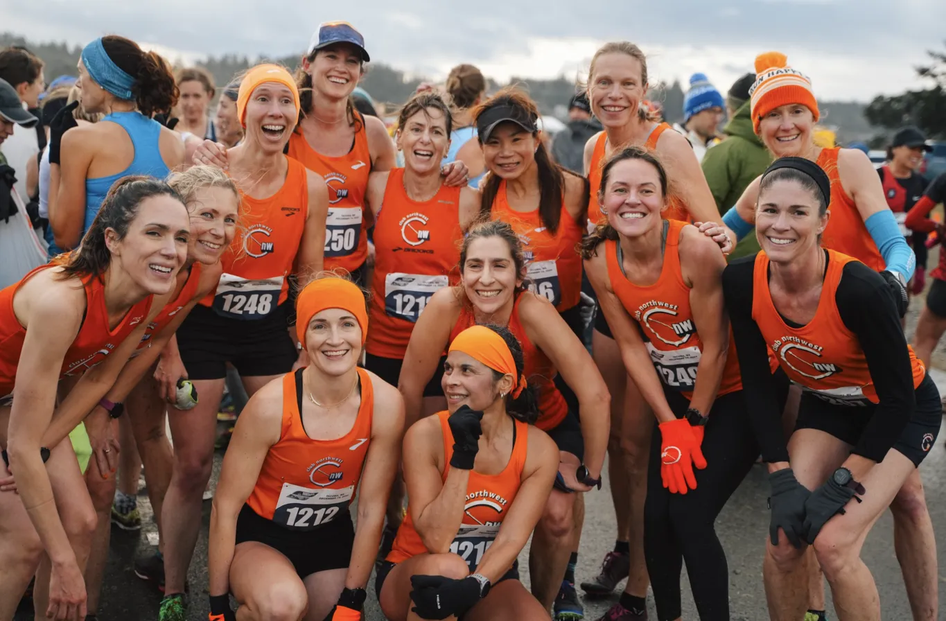 A group of runners in bright orange Northwest running team jerseys gather for a pre-race photo at Magnuson Park, sporting winter running gear and enthusiastic smiles against an overcast Seattle sky.