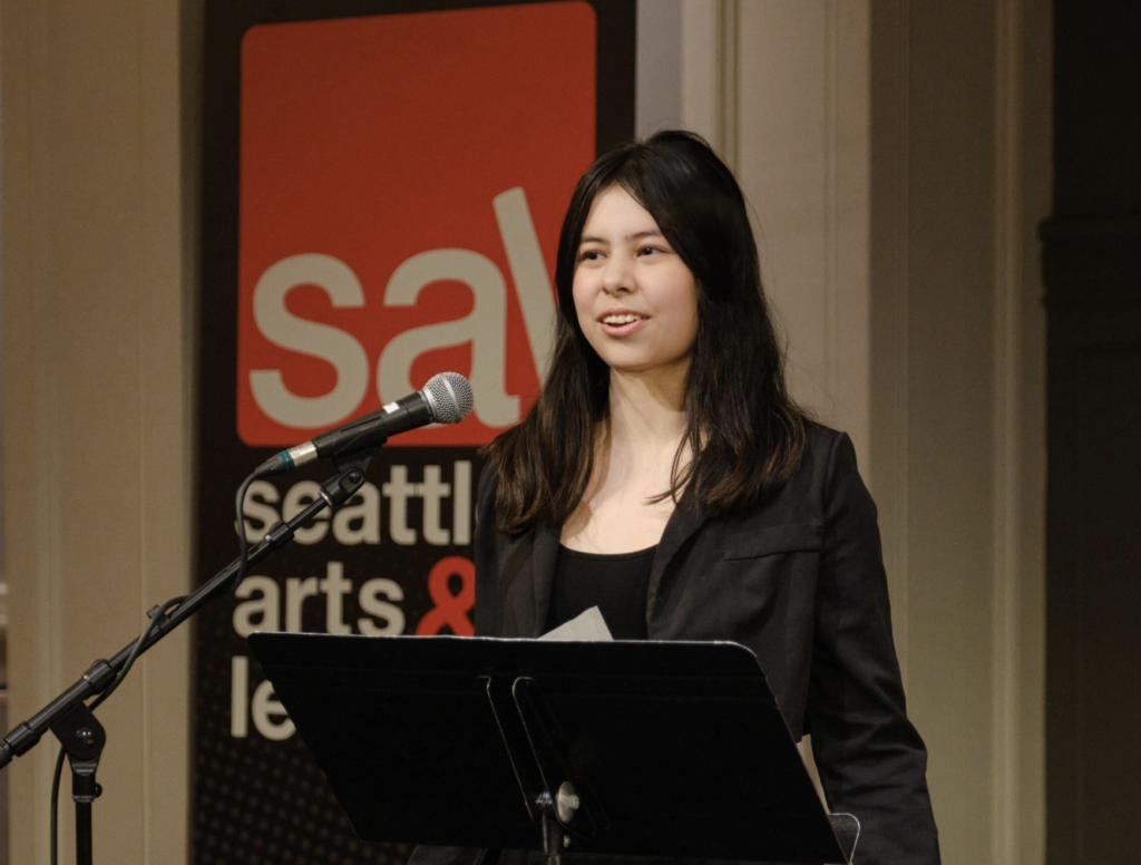 A speaker stands at a podium with a microphone against a backdrop showing the red and white SAL (Seattle Arts & Lectures) logo.