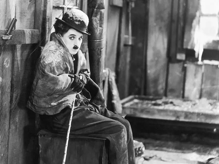 Black and white still from Charlie Chaplin's 'The Gold Rush' showing Chaplin as his famous Tramp character sitting on a ledge in tattered clothes and his signature bowler hat, holding a cane and looking directly at the camera with his characteristic expression.