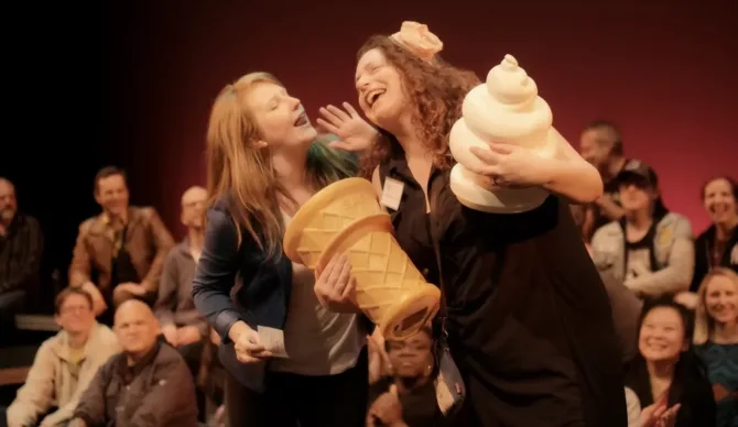 Two women on stage laughing heartily while holding large prop ice cream cones - one yellow waffle cone and one white soft-serve. They're performing in front of an audience visible in the background against a dark red stage lighting.