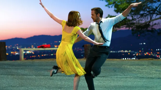 Two dancers in an iconic scene from La La Land perform against a twilight Los Angeles skyline, with one wearing a flowing yellow dress and the other in business attire as they strike a symmetrical pose with outstretched arms.