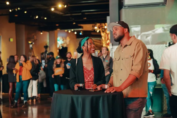 nside MoPOP's warmly lit exhibition space, guests gather around a cocktail table draped in black cloth during an evening event. Two people share a moment of engaging conversation, with ambient lighting creating an inviting atmosphere for cultural discussion and community connection.