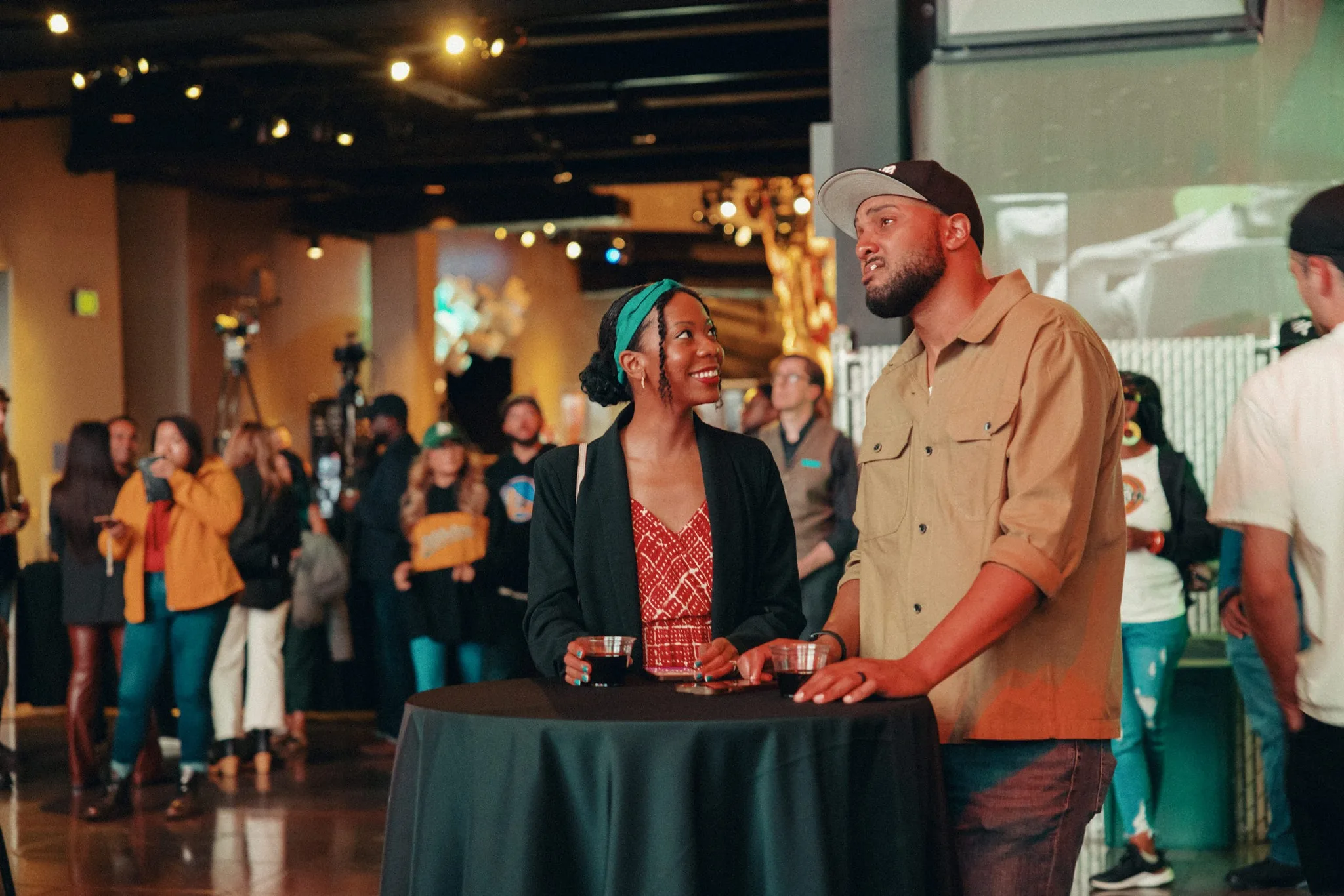 nside MoPOP's warmly lit exhibition space, guests gather around a cocktail table draped in black cloth during an evening event. Two people share a moment of engaging conversation, with ambient lighting creating an inviting atmosphere for cultural discussion and community connection.