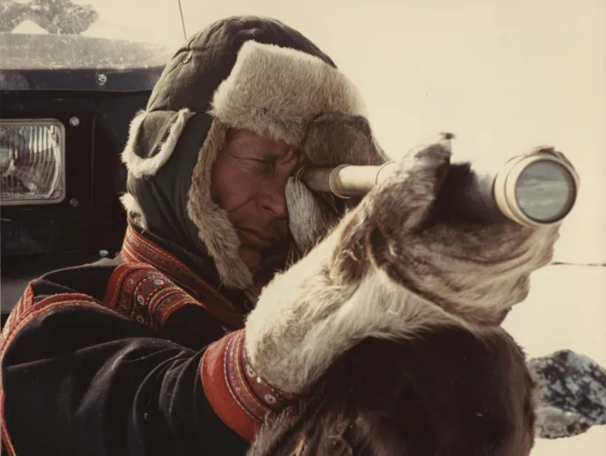 A person in traditional Sámi winter clothing with a fur-lined hat looks through a telescope or camera lens, photographed against a bright winter sky. The individual wears a dark coat with traditional red embroidered trim.