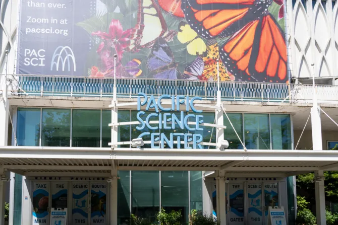 The Pacific Science Center building exterior features its iconic blue neon signage against a glass facade, topped by a large colorful butterfly mural banner. White architectural lattice work frames the entrance, with wayfinding signs for "Main" and "Gates" visible at ground level.