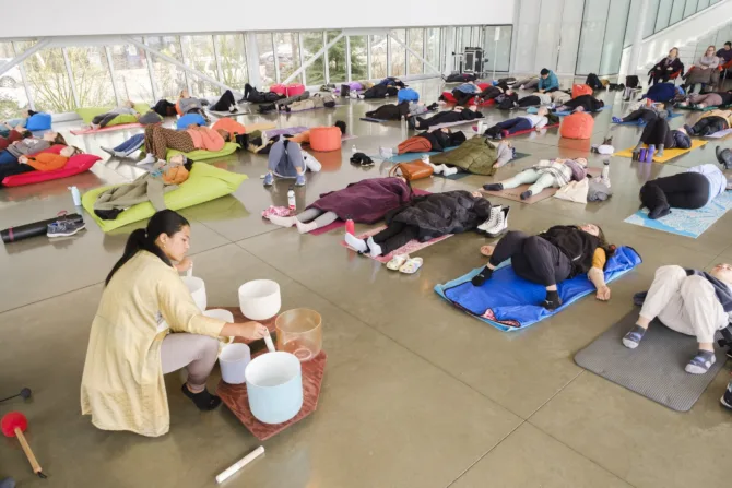 A person in a yellow sweater sits cross-legged beside crystal sound bowls while participants rest on colorful yoga mats in the light-filled PACCAR Pavilion, with Seattle's waterfront visible through floor-to-ceiling windows.