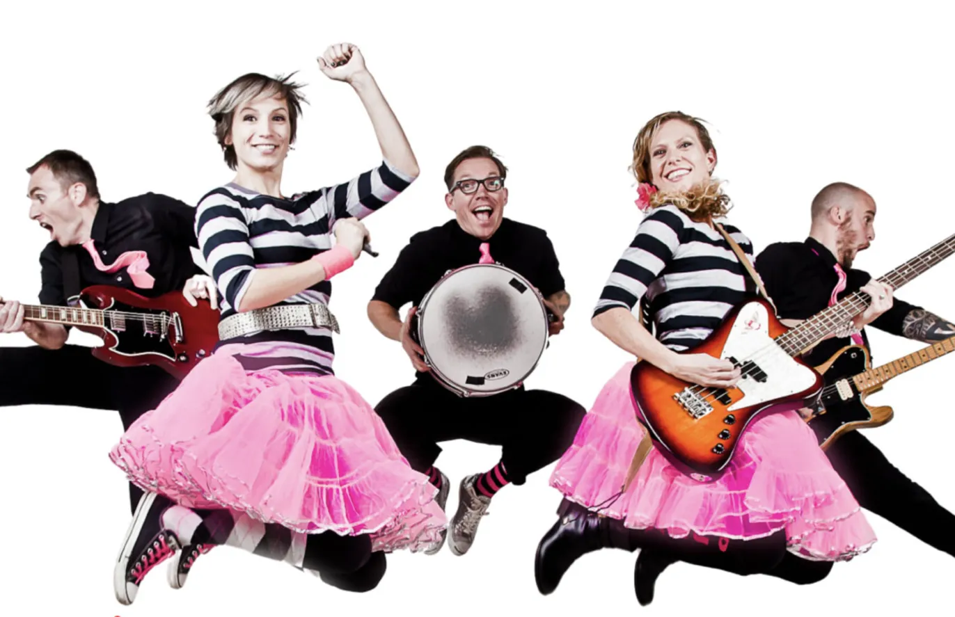 Five energetic musicians from The Not-Its! leap simultaneously during a performance - two guitarists and a drummer in black, with two vocalists in striped shirts and pink tutus, all with joyful expressions.