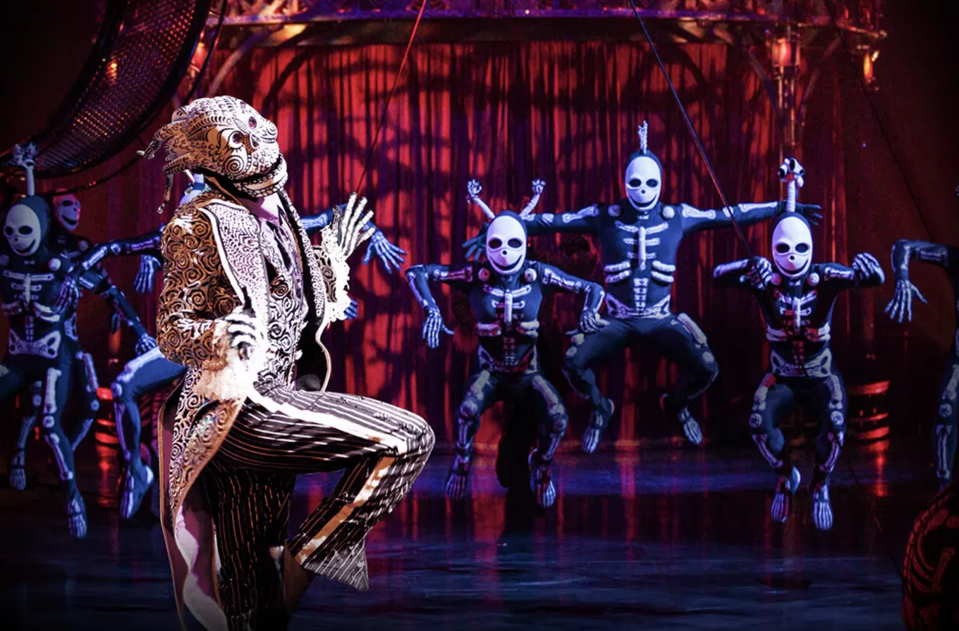 A dramatic scene from Cirque du Soleil's KOOZA shows a performer in an ornate gold and white costume against a red curtain backdrop, while skeleton-suited acrobats leap through the air behind them.