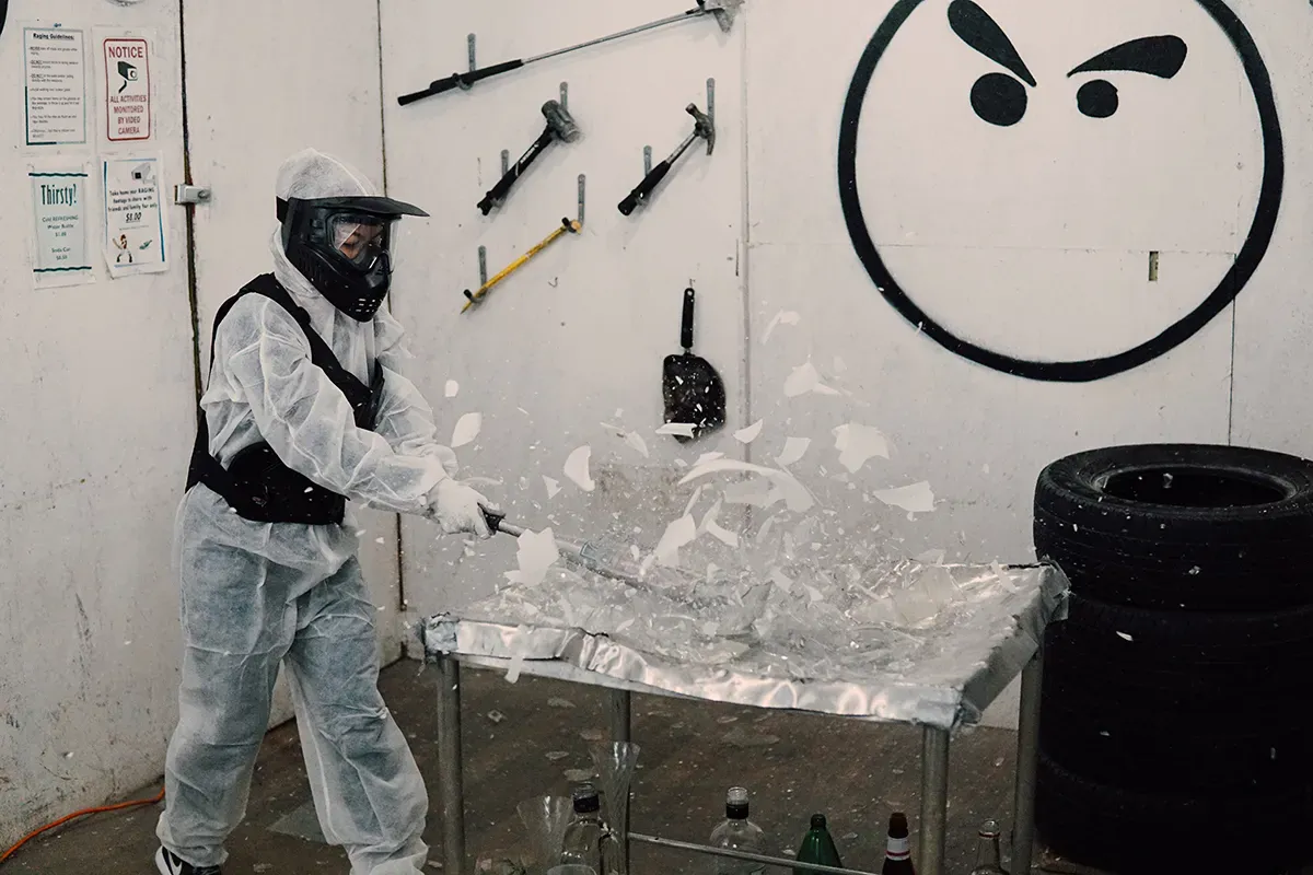 Person in white protective suit and black face mask swings a bat at items on a metal table, creating dramatic spray of shattered debris. Large black circular logo visible on white wall behind them, with tool holders displaying various smashing implements mounted nearby.