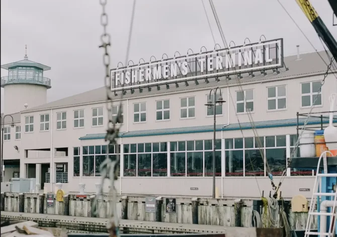 The Fishermen's Terminal building in Seattle on a gray day, with its iconic sign mounted on the white facade. A distinctive teal watchtower stands at one end, and the building's long row of windows reflects the marina. Weathered dock pilings and marine equipment are visible in the foreground.