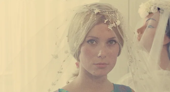 A close-up fashion photo showing a bridal look, featuring a delicate vintage-style veil adorned with small beaded details and floral embellishments. The veil has a soft, ethereal quality in warm lighting. The person is wearing subtle makeup and appears in a dreamy, romantic style reminiscent of 1960s fashion photography.