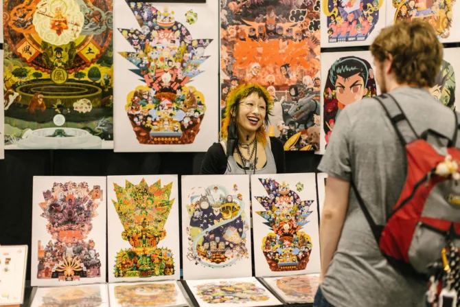 An artist in Artist Alley smiles while talking to a convention-goer from behind their booth.