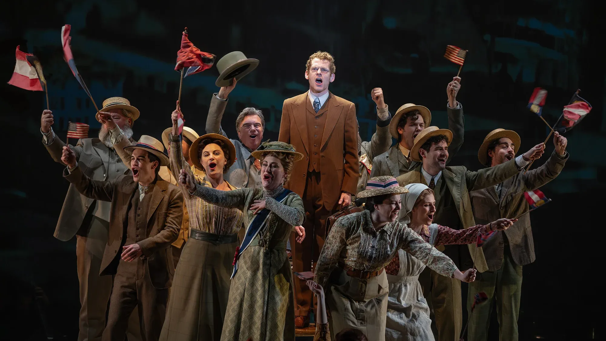 A crowd of people singing and waving flags gather around a person standing on a chair, spotlighted onstage.