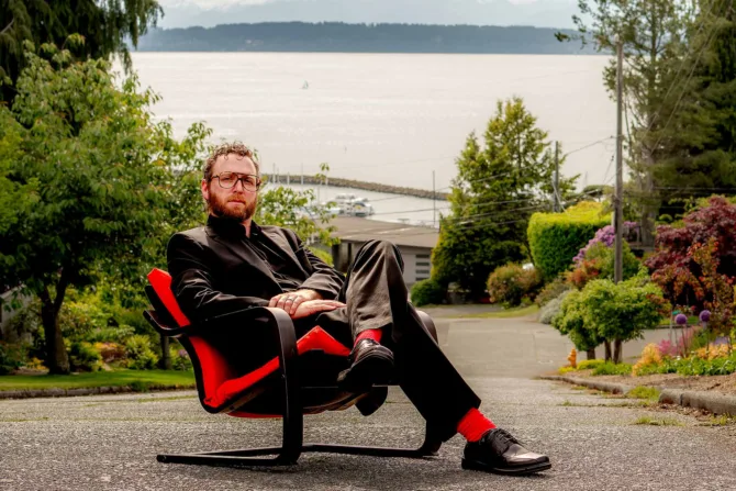 Brian Pucheu, as Ingvar Kamprad, sits in his IKEA chair in front of mountains.